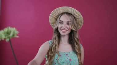 Bir çiçekle kameraya poz veren dalgalı saçlı neşeli genç bir kadın, onu veriyor ve mutlu bir şekilde gülümsüyor. Pozitif duygular, gerçek neşe, iyi hissetmek. Şık çiçekli görünüş, modaya uygun şapka. Renkli arka plan, kadın portresi