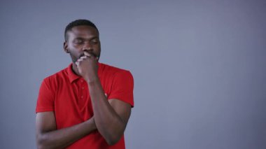 Parlak kırmızı stil polo tişörtlü yakışıklı Afro-Amerikalı adam çenesine el ele tutuşmuş, gri arka planı düşlüyor. Sorunlar, sorunlar, planlama. Erkek portresi