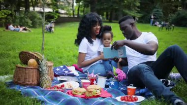 Şirin Afrikalı bir aile piknik battaniyesinde oturup fotoğraflara bakıyor. Yakışıklı genç adam karısına ve kızına resimlerini gösteriyor. Piknik. Açık havada. Yaz zamanı. Sıcak hava.
