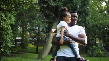 Hoş görünümlü, sıradan giysiler içinde, küçük kızını elinde tutan Afrikalı genç bir adam. Güzel bir çocuk, pipetle annesine bakıyor. Açık havada. Yaz zamanı..