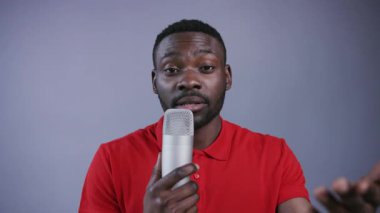 Kırmızı polo tişörtlü neşeli Afro-Amerikalı adam mikrofon kullanıyor ve gri arka planda konuşuyor. Stand-up komedyeni, şaka yapıyor, eğleniyor, kayıt yapıyor. Erkek portresi, yakın görünüm