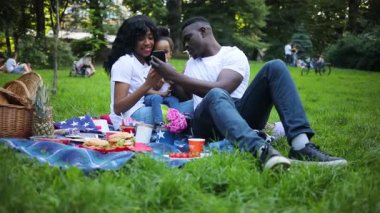 Sevimli aşık bir çift ve piknik battaniyesinde yan yana oturan küçük bir kız. Siyah akıllı telefonu elinde tutan genç bir Afrikalı. Fotoğraflar gösteriyor, gülümsüyor..