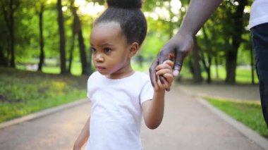 Parkta yürüyen ve genç babasıyla el ele tutuşan şirin saçlı Afrikalı bir bayan. Baba ve kızı. Güneş ışığı. Yaz zamanı. Gündüz vakti. Park.