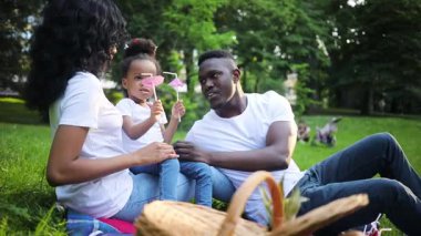 Küçük tatlı Afrikalı kız pipet kokteyli oynuyor. Piknik battaniyesinde oturan mutlu bir aile. Çocuk annesinin kucağında oturuyor. Açık havada. Gündüz vakti. Yaz zamanı. Güneş ışığı.