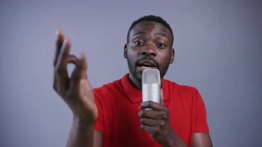 Yakışıklı Afro-Amerikalı bir adam doğrudan kameraya bakar, mikrofonla konuşur, gri arka planda hareketli hareketler yapar. Sunum, vaiz, stand-up, tartışma kulübü, tartışma. Erkek portresi, yakın görünüm