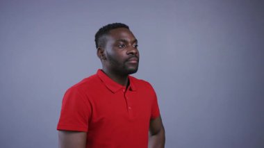 Parlak kırmızı gömlekli yakışıklı Afro-Amerikalı adam sabit duruyor, sonra doğrudan kameraya bakıyor ve gri arka planda baştan çıkarıcı bir şekilde gülümsüyor. Erkek güzeli. Erkek portresi, yakın görünüm