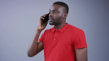 Parlak kırmızı polo tişörtlü yakışıklı Afro-Amerikalı adam telefona cevap veriyor ve gri arka planda gülümsüyor. Akıllı telefon, cihazlar, iletişim. Erkek portresi, yakın görünüm