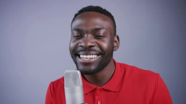 Adam şarkı söylüyor. Genç Afro-Amerikalı şarkıcı mutlu bir gülümsemeyle mikrofonun yanında duruyor, şarkı söylüyor ve gri arka planda dans hareketleri yapıyor. Kayıt, stüdyo, karaoke. Erkek portresi, yakın görünüm