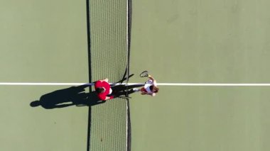 Bir erkek ve kadının tenis maçına başlarken yüksek açılı çekimleri, ağın yanında buluşmaları, birbirlerine beşlik çakmaları, pozisyonlarında yürümeleri, topu atmaları, erkeklerin topu atmaları, kadınların kaçırması. Spor için içeri gir..