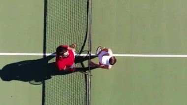 Yüksek açılı bir atış, tenis oyuncularının ağın yanında buluşması, beşlik çakması, pozisyonlarının üzerinde yürümeleri. Maçın başlangıcı, tenis oynamak, spor yapmak, aktif yaşam tarzı. Helikopter vuruldu.