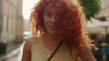 Kızıl kıvırcık saçlı bir kadın sokakta yağmurda yürüyor kameraya bakıyor mutlu bir gülümseme takınıyor güzel bir moda su silueti yaz yüzü yalnız durun