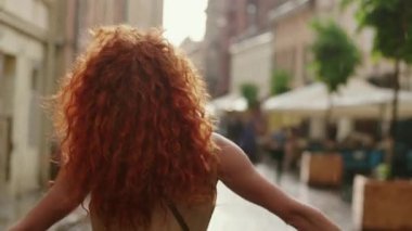 Şık, kıvırcık saçlı, kırmızı kıvırcık saçlı bir kadın sokakta yağmurda yürüyor, kameraya bakıyor, gülümsüyor, gülümsüyor, modayla ilgili tatlı bir gülücük yaz silueti.