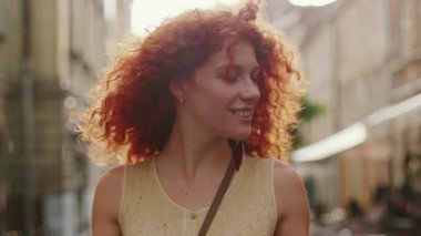 Kırmızı kıvırcık saçlı, gülümseyen genç bir kadın yağmur altında yürüyor. Kameraya bak. Mutlu moda su siluetini çeviriyor.