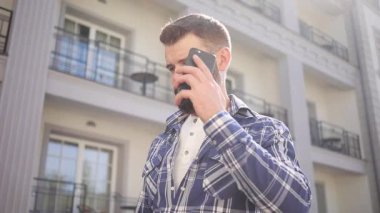 Sakallı, kareli tişörtlü, parlak güneş ışığı altında vahşi bir adamın düşük açılı görüntüsü. Bir telefon alır, telefona cevap verir. Sohbet, olumlu duygular, iyi hissetmek. Mutluluk. Erkek portresi