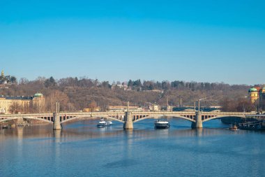 Prag 'ın tarihi kemerli köprüsü olan manzaralı Vltava Nehri ve ağaçlarla kaplı bir tepenin üzerindeki uzak Petrin Gözcü Kulesi berrak, güneşli mavi bir gökyüzünün altında. Prag, Çek Cumhuriyeti - 19 Şubat 2025.
