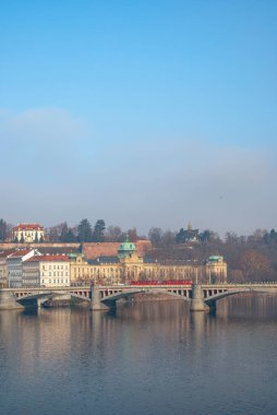 Prag 'ın Vltava Nehri' nin tarihi kemerli köprüsü ile panoramik manzarası, nehir kenarlarında geleneksel mimari ve uzak bir tepe sergileniyor. Prag, Çek Cumhuriyeti - 24 Şubat 2025.