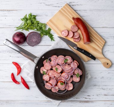 Uncooked calabrese sausage slices in a pan with onion, pepper and herbs over white wooden table.