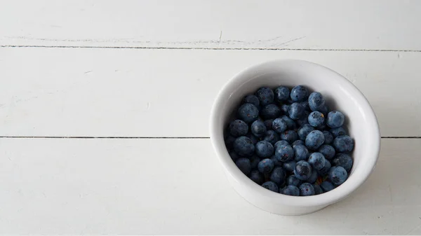 stock image  Flat lay view of a small white ceramic bowl filled with fresh blueberries. Placed on top of a wooden table                              