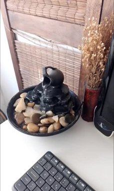 Close view of a decorative zen water fountain next to a keyboard, a monitor and dry flowers decorative bouquet. on top of a white wooden desk