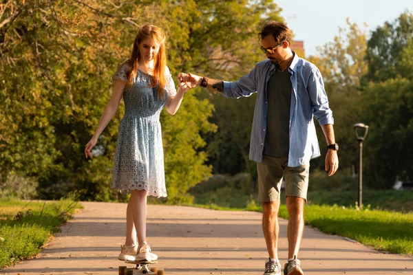 Kucaklayan ve kız arkadaşına asfalt yolda ağaçlar arasında uzun tahta üstünde yardım eden genç adam.
