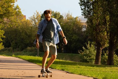 Yeşil ağaçlar arasındaki boş asfalt yol boyunca uzun tahta üzerinde giden genç adamın ön görüntüsü.