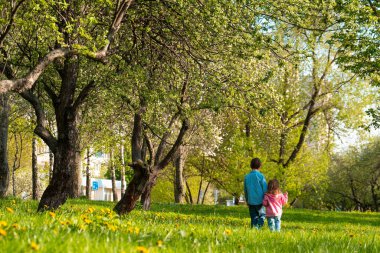 Kardeşiyle birlikte yeşil parktaki ağaçların arasında el ele tutuşan küçük kızın arka görüntüsü.