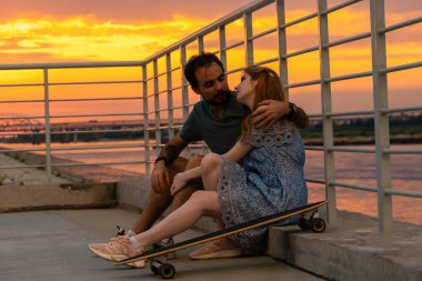 Romantik genç çiftin gün batımına karşı sette otururken birbirlerine baktıkları yan manzara.