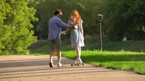 木の間のアスファルトのウォークウェイでロングボードに乗っているガールフレンドを抱擁し 助ける若い男のバックビュー — ストック動画
