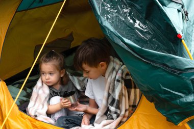 Küçük bir çocuk ve ekoseye sarılı küçük bir kız kırsaldaki bir çadırda oturmuş sıcak çay içiyorlar. Kamp ya da mültecilik kavramı