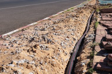 Dig a trench to lay cables in a conduit. Dig a long trench to place the electrical wiring in a black plastic conduit in the ground on the side of the paved road with copy space. Selective focus clipart