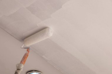 Paten tavanı boyuyor. Uzun saplı bir paten fırçasının yakın çekimi yenilenmiş bir ev veya binada alçı taşı tavanına beyaz bir astar çizer..