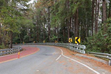 Curve road sign on down hill. Black arrow sign on yellow background on pole and silver metal barricade warning to drive downhill in forest with caution on green tree background and selective focus.