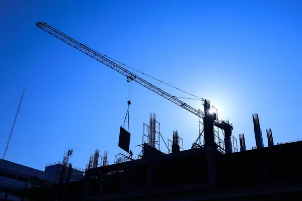 Silhouette of a crane on a building. Industrial construction crane lifting steel sheets on tall building under construction on sunrise background in bottom view with copy space with selective focus.