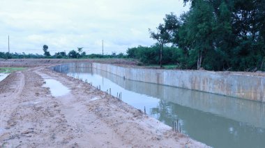 Beton sulama kanalı. Tarım drenajı için sağda yeşil ağaçları olan yeni inşa edilmiş bir kanal ve bulutlu arka planda Tayland 'da sel koruması. Seçici odak