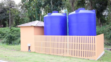 Two Plastic Water Storage Tanks. Large Blue Plastic Water Storage Tanks in a Barn with an Outdoor Water Pump for Water Storage on Green Trees Background with Copy Space. Selective Focus clipart