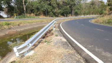 Virajda korkuluk var. W-Beam 'in yakın plan görüntüleri veya kavisli yan direğe kurulmuş metal çitler, kanal ve yeşil ağaçların seçici odaklı arka planlarına karşı araba kazalarını önlüyor.