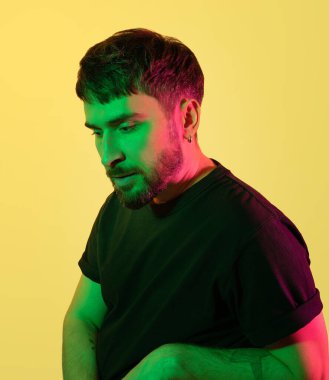 Sanatsal yaratıcı portre. Sarı arka planda yeşil, pembe neon ışıklarla bakan dalgın yakışıklı bir adam. Modern fotoğraf. Kulağında küpe olan güzel sakallı orta yaşlı adam. Boşluğu kopyala.