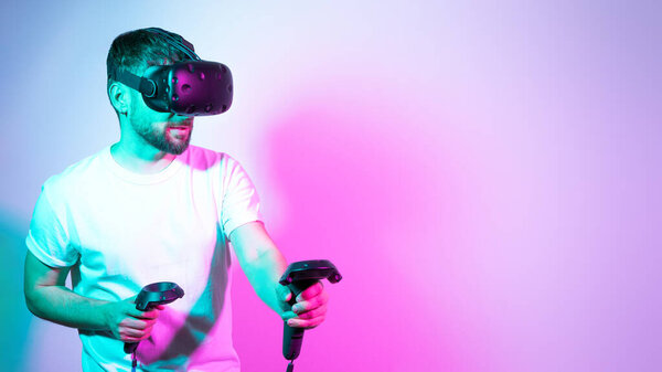 Concept modern hobby. Bearded millennial man in VR glasses plays in simulation figth and using controllers in gradient neon background. interactive online game in a virtual world. Copy space, banner.