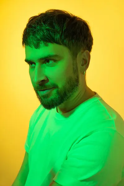 Creative portrait. Smiling man with a sly look in green neon lights on yellow background. Modern photo handsome bearded middle age man with earring in the ear.