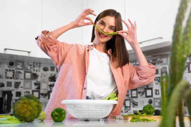 Mutlu, genç, güzel, neşeli kız ev mutfağında yeşil fasulyeden bıyık yapıyor ve gülümsüyor. Sağlıklı beslenme ve vejetaryen yaşam tarzı kavramı. Yeşil sebze. Gerçek duygular.