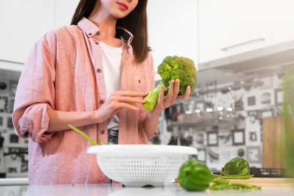 A Young woman in oversize pink shirt stands in the home kitchen, holding green broccoli in hands, going to cook a vegetarian diet food. The concept of a healthy nutrition, and vegan lifestyle.