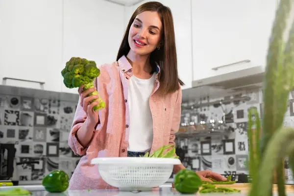 A Young woman stands in the home kitchen, holding broccoli in hands, going to cook a vegetarian diet salad. The concept of a healthy nutrition and vegan lifestyle. Healthy food for detox. Copy space.