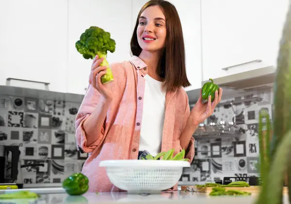 A Young smiling woman holding broccoli, bell pepper in hands. The concept of a healthy nutrition and preparing vegetarian food at home kitchen. Healthy food for detox. Vegan lifestyle. Copy space.