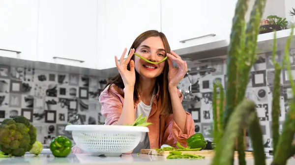 Happy Young beautiful cheerful girl making mustache from green bean on face at home kitchen and smiles. The concept of a healthy nutrition and vegan lifestyle. Healthy food for detox. Copy space.