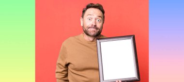 Orta yaşlı bir adam omuz silkiyor, kafası karışık ve boş bir çerçeve ile kararsız.