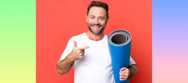 Orta yaşlı bir adam neşeli bir şekilde gülümsüyor, mutlu hissediyor ve bir tarafı işaret ediyor. Yoga mattıyla. fitness konsepti