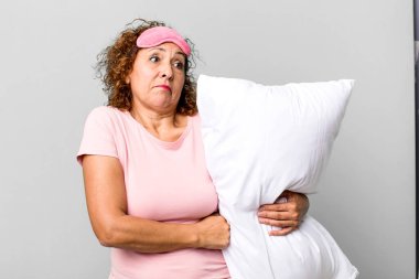 Güzel orta yaşlı bir kadın omuz silkiyor, kafası karışık ve kararsız. Pijama geceliği ve yastık giyiyor.