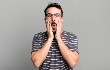 Şok olmuş ve korkmuş hissediyorum, ağzı açık ve elleri yanaklarında korkmuş görünüyor.