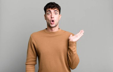 Adam şaşırmış ve şoka uğramış görünüyor, çenesi düşmüş ve eli açık bir objeyi tutuyor.