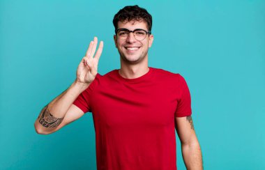 Gülümsüyor ve dost canlısı görünüyor. Üç ya da üç numarayı gösteriyor. Elleri önde, geriye doğru sayıyor.